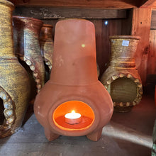 A terracotta colored mini chiminea candleholder with a candle burning inside it, placed on a wooden shelf with other ceramic pots in the background.