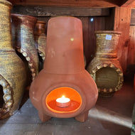 A terracotta colored mini chiminea candleholder with a candle burning inside it, placed on a wooden shelf with other ceramic pots in the background.